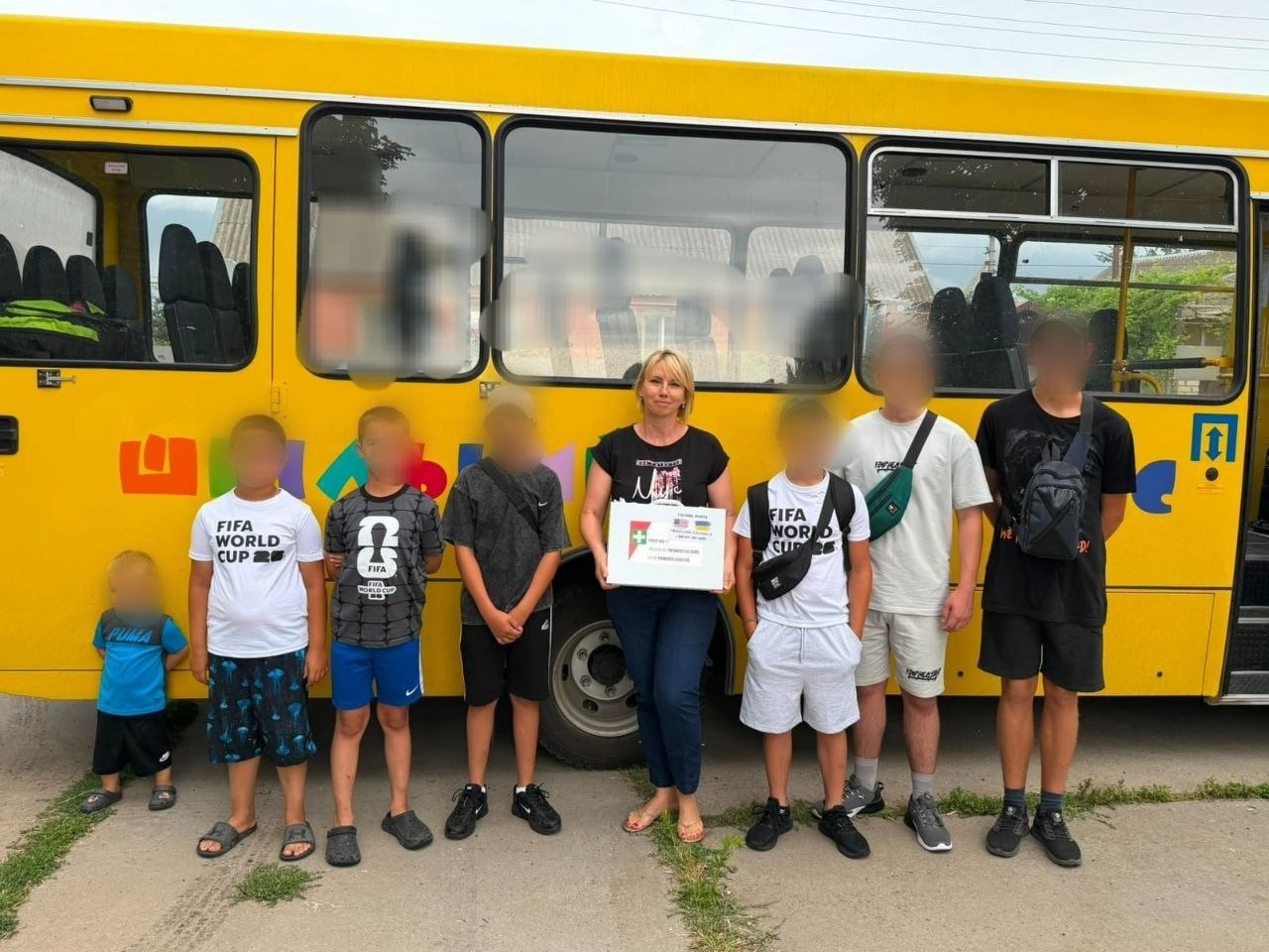 Волонтерка та група хлопчиків і підлітків із розмитими (заблуреними) з міркувань безпеки обличчями стоять у ряд перед жовтим автобусом, тримаючи в руках подяку від американських благодійників «Мандрівні полковники», які разом із Голопристанською громадою передали чергову партію допомоги для Сил оборони.