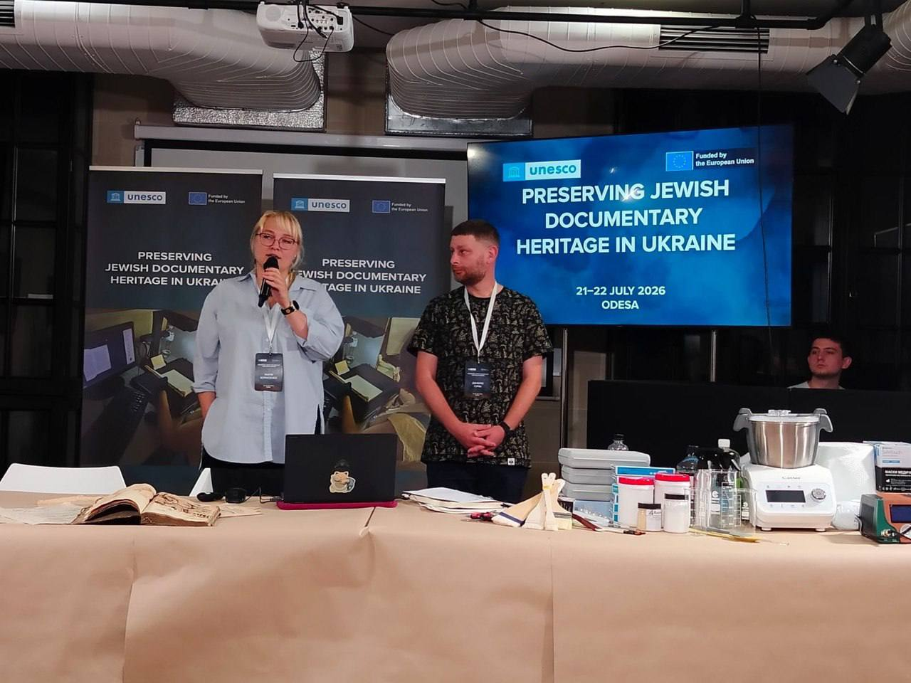 Спікерка виступає з мікрофоном, а поруч стоїть її колега біля довгого столу з реставраційним обладнанням і старовинними книгами під час тренінгу в Одесі на тлі великого екрана та банерів ЮНЕСКО з написом «Preserving Jewish Documentary Heritage in Ukraine».