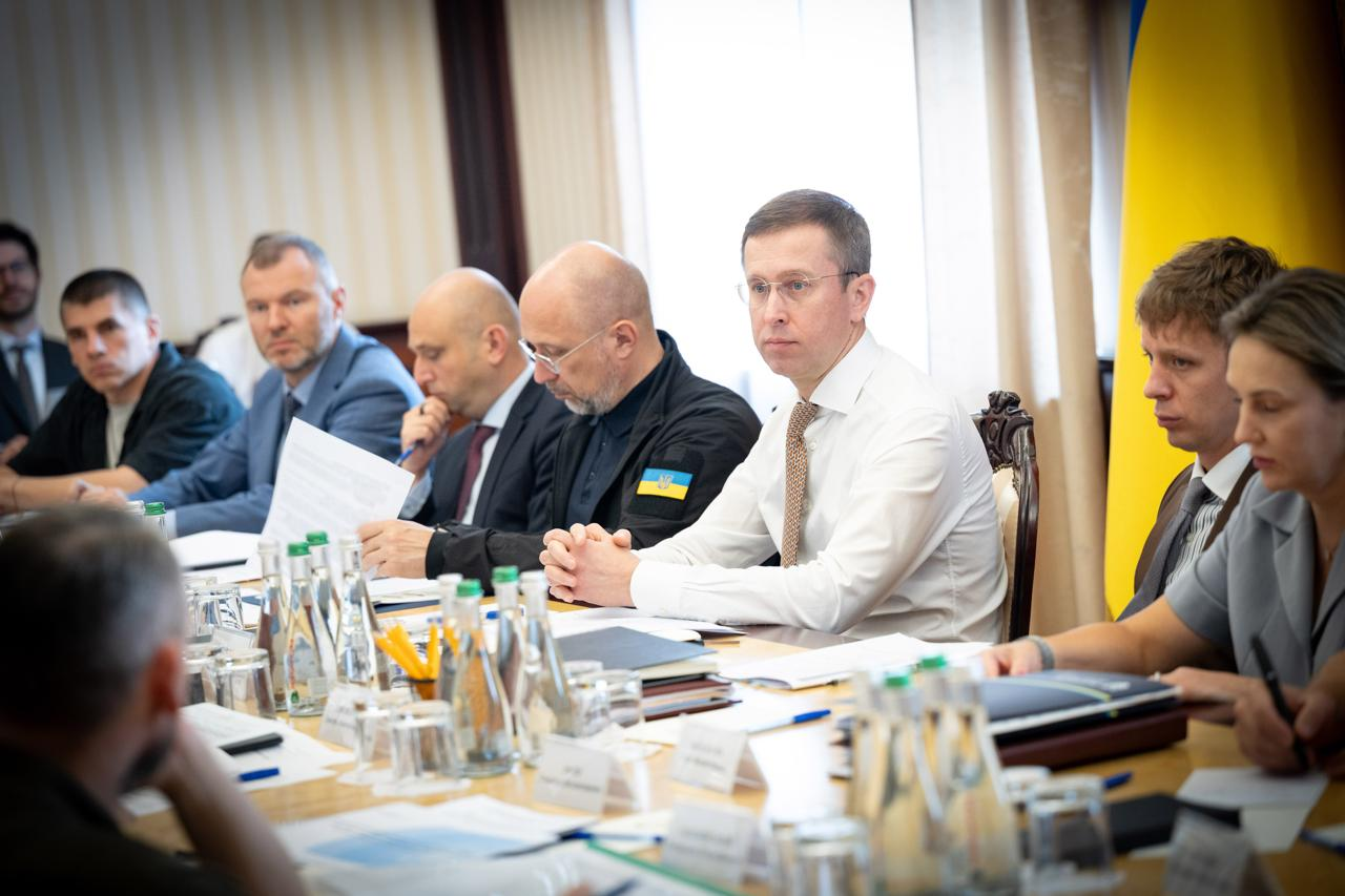 Прем'єр-міністр Сергій Корецький (у білій сорочці та краватці) та Денис Шмигаль (у чорній тактичній куртці з прапором України) сидять за столом президії в оточенні урядовців на нараді щодо виконання планів стійкості до зимового періоду. На передньому плані видно документи та пляшки з водою.