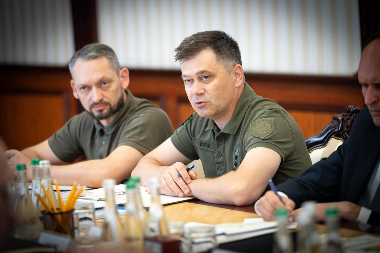 Голова Держспецзв'язку Олександр Потій (у центрі, в поло кольору хакі з іменним патчем) виступає на офіційному засіданні за робочим столом. Поруч із ним сидять інші посадовці у військовій формі та ділових костюмах. На передньому плані видно канцелярію та пляшки з водою.