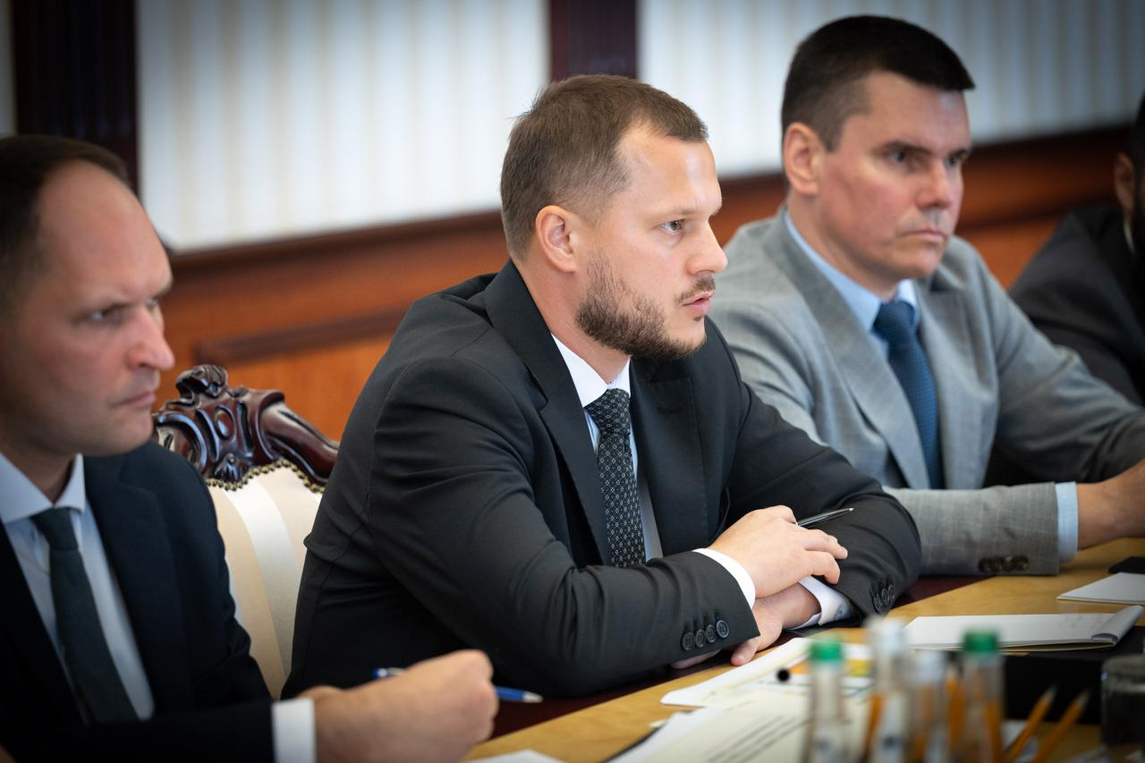 Посадовець у темному костюмі та краватці (у центрі) виступає на офіційному засіданні за робочим столом. Ліворуч і праворуч від нього сидять інші учасники наради у ділових костюмах, які слухають доповідача.