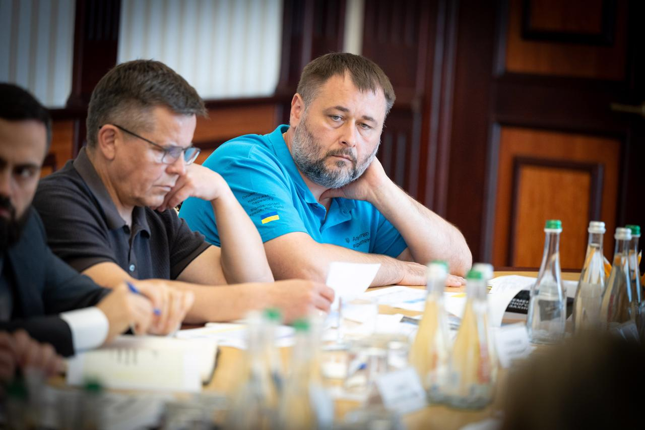Посадовець у блакитному поло із прапором України на рукаві сидить за столом президії поруч із колегами, підперши рукою підборіддя, та уважно слухає виступи під час урядової наради. На передньому плані на столі стоять пляшки з водою та розкладені документи.