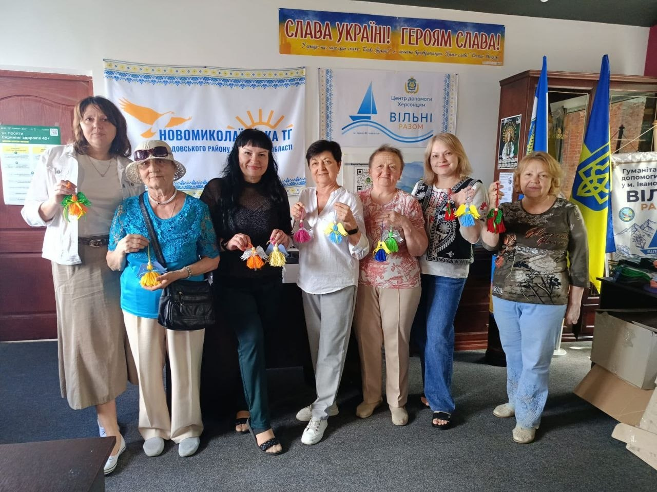 Група переселенок із Херсонщини стоять у ряд у кімнаті координаційного центру «Вільні разом» в Івано-Франківську. Жінки посміхаються та тримають у руках виготовлені з кольорових ниток підвіски у вигляді янголів. На задньому плані розміщені банери з патріотичними гаслами, назвами організацій та державні прапори України.