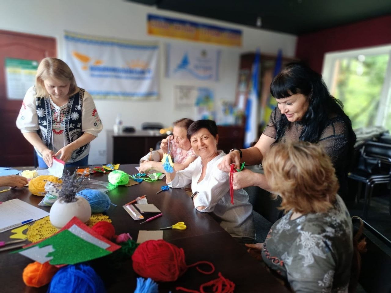Переселенки з Херсонщини виготовляють нитяні підвіски у вигляді янголів на майстер-класі в центрі «Вільні разом» в Івано-Франківську. Жінки з посмішками сидять за столом із кольоровою пряжею, ножицями та заготовками. Одна з жінок (праворуч) стоїть і допомагає учасницям із плетінням, а жінка у вишиванці ліворуч готує матеріали. На задньому плані видно патріотичну символіку.