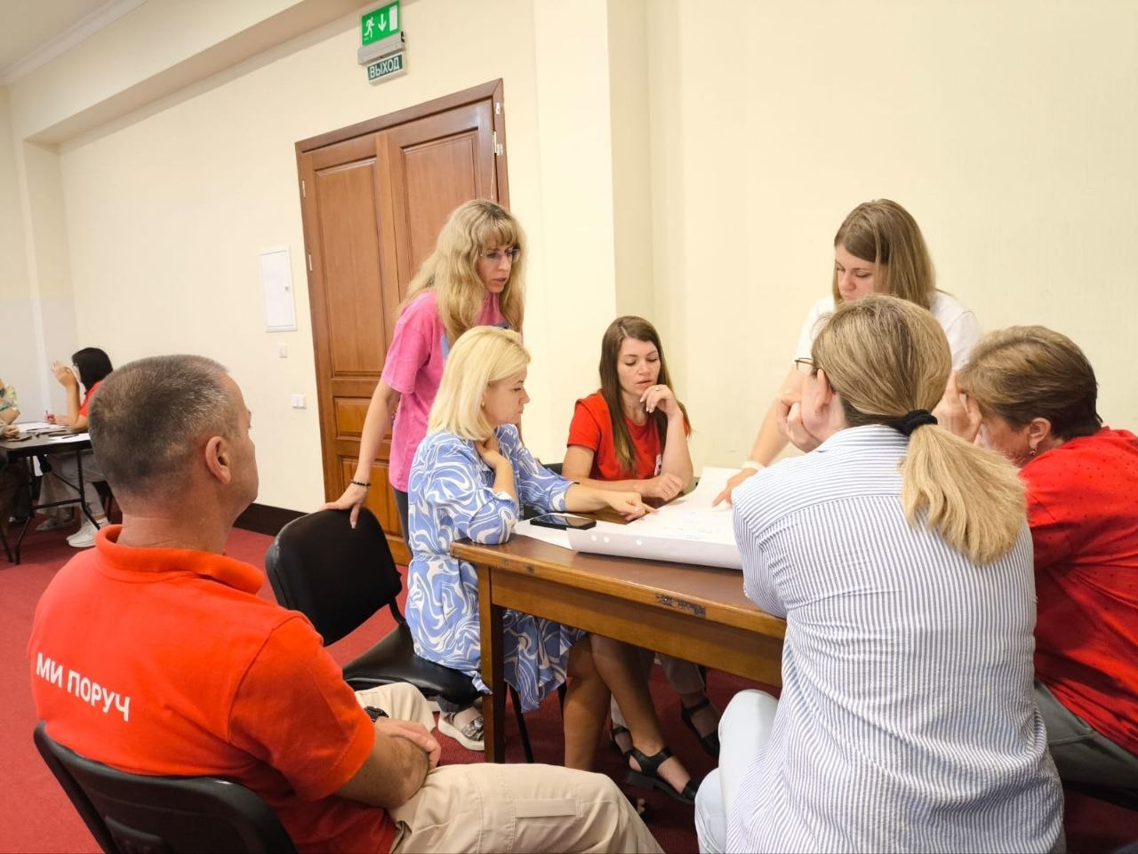Координаторка центру «Вільні разом» у Житомирі Алєся Алєксєєва (стоїть зліва у рожевій футболці) разом з іншими учасниками та волонтерами Українського Червоного Хреста аналізує навчальні матеріали під час тренінгу з волонтерського менеджменту в Одесі. Група людей сидить за великим дерев'яним столом у кабінеті та зосереджено обговорює написи на великому паперовому ватмані. На передньому плані ліворуч сидітиме чоловік у червоній фірмовій футболці з написом «МИ ПОРУЧ».