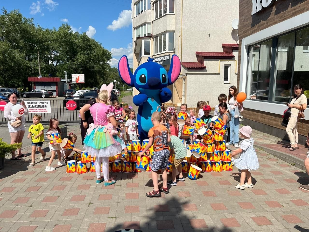 Діти-переселенці з Херсонщини граються на святі від центру «Вільні разом» та «Royal Hamburger» у Хмельницькому. Малеча разом з аніматоркою у рожево-бірюзовому костюмі та великою ростовою лялькою Стіча збирає вежу з яскравих різнокольорових відерець на тротуарі. Ліворуч на фоні видно паркан із табличкою «ОСББ "Світанок"» та знак «STOP».