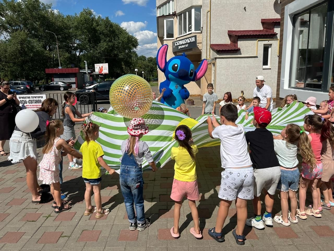 Діти-переселенці з Херсонщини граються на святі від центру «Вільні разом» та «Royal Hamburger» у Хмельницькому. Хлопчики та дівчатка тримають за краї велике зелено-біле смугасте полотно, підкидаючи на ньому велику прозору диско-кулю. На задньому плані дітей розважає велика ростова лялька синього Стіча, а ліворуч видно паркан із табличкою ОСББ та білу повітряну кульку.