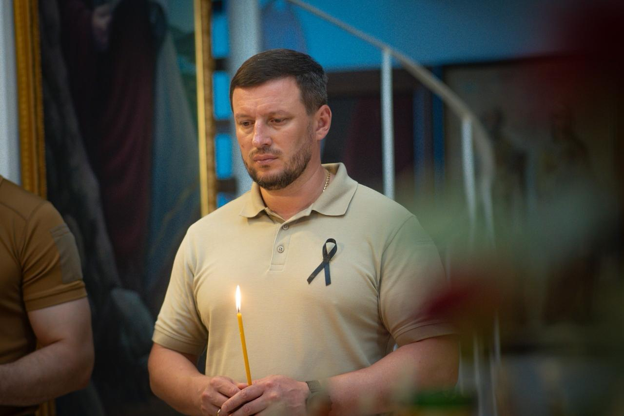 Начальник Херсонської ОВА Олександр Прокудін у тактичному поло з чорною траурною стрічкою стоїть у храмі із запаленою свічкою під час вшанування пам’яті жертв теракту в Оленівці та всіх страчених і загиблих у російському полоні. У нього сумний і зосереджений вираз обличчя, погляд спрямований донизу. На розмитому фоні видно церковні ікони.