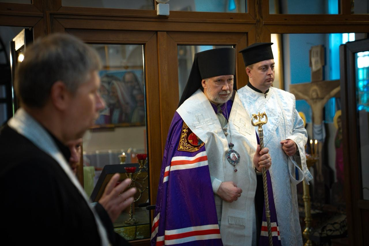 Священнослужитель у фіолетовому та білому церковному вбранні з панагією на грудях тримає посох під час панахиди у храмі за загиблими захисниками України. Праворуч за ним стоїть помічник у світлій ризі, а ліворуч на передньому плані видно профілі інших священників, які беруть участь у службі.
