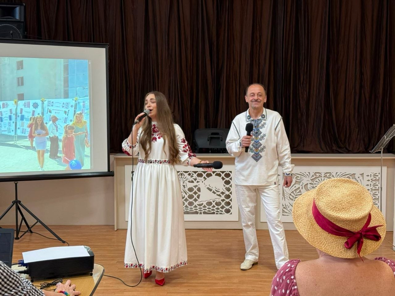 Виступ артистів на відкритті виставки «Херсонщина вишивана» в Одесі. У центрі сцени дівчина у довгій білій сукні з червоною вишивкою співає в мікрофон. Праворуч від неї стоїть чоловік у білій вишиванці та білих штанях із мікрофоном у руках. Ліворуч на сцені розміщено великий проєкційний екран із фотографією, а на передньому плані праворуч видно глядачку в солом'яному капелюсі.