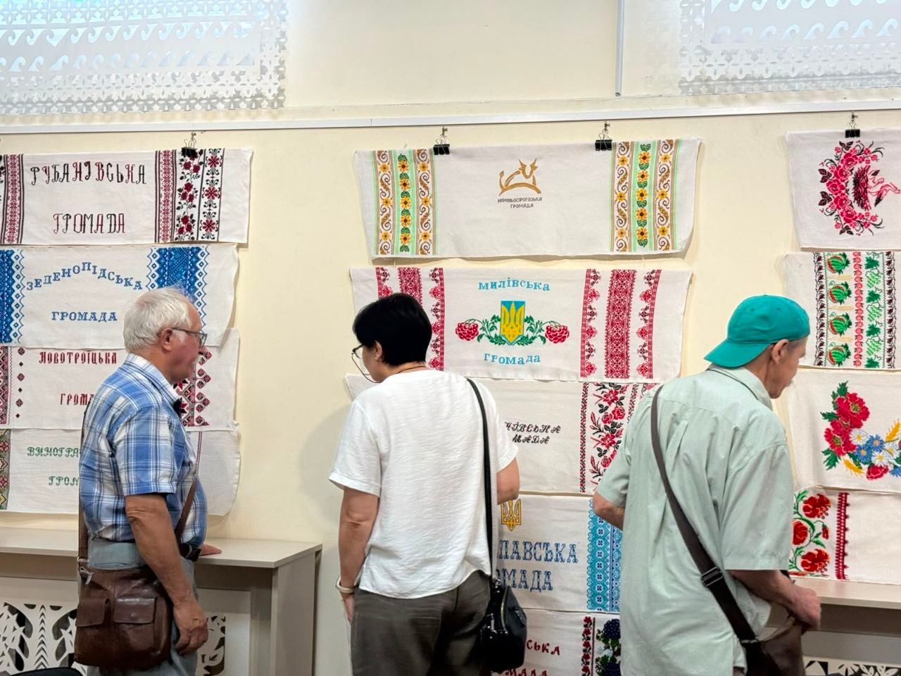 Відвідувачі розглядають вишиті рушники громад Херсонської області на виставці «Херсонщина вишивана» в Одесі. На стіні представлені роботи з написом «Рубанівська громада», «Зеленопідська громада», «Милівська громада» та інших ТГ. Експозицію вивчають сивий чоловік у картатій сорочці (ліворуч), жінка у білій футболці (у центрі) та чоловік у зеленій кепці (праворуч).