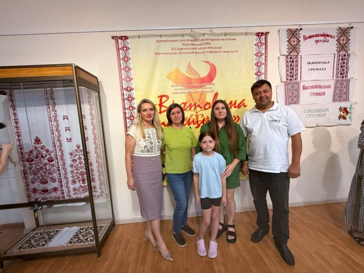 Група з п'яти людей (три жінки, чоловік та дівчинка) фотографується на виставці «Херсонщина вишивана» в Одесі. Вони стоять на тлі банера проєкту «Врятована спадщина» та розвішаних на стіні рушників із назвами «Великоолександрівська громада», «Іванівська громада», «Високопільська громада» та «Скадовська громада». Ліворуч розміщена скляна вітрина з автентичним вишитим текстилем.