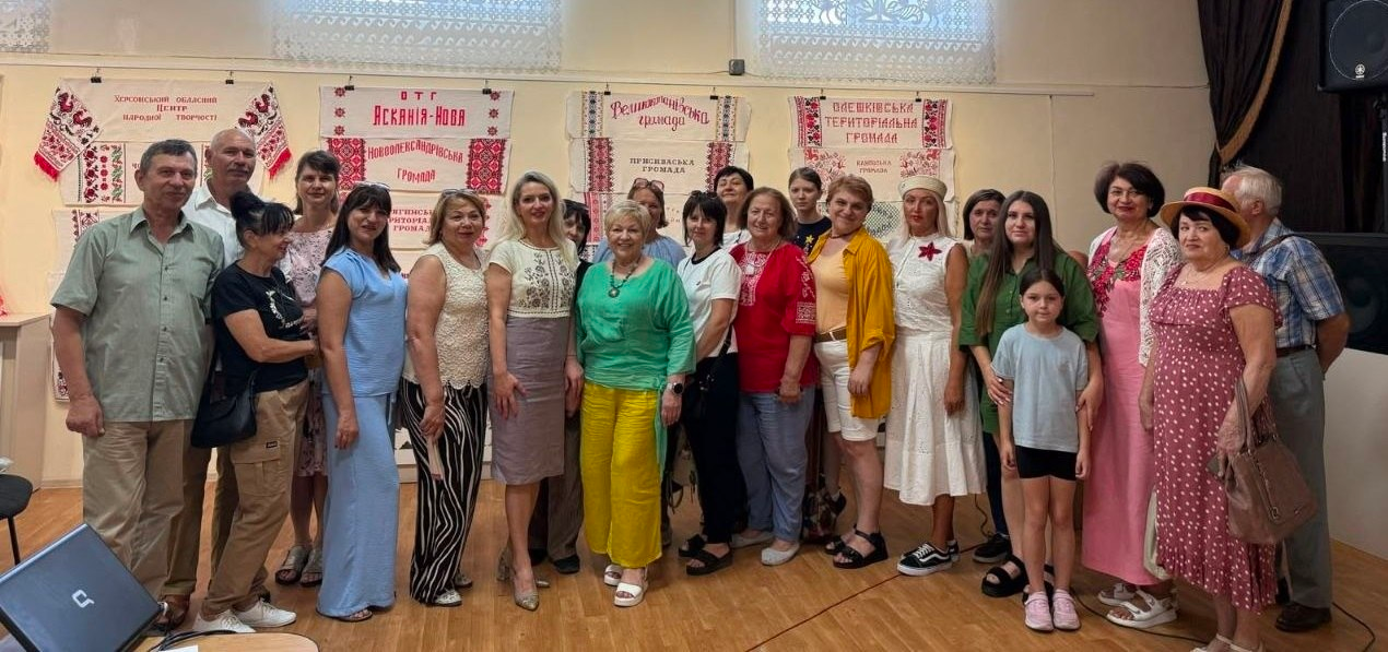 Велика група відвідувачів та організаторів фотографується разом у залі на виставці «Херсонщина вишивана» в Одесі. Чоловіки, жінки та діти стоять у ряд на тлі стіни, де розвішані врятовані вишиті рушники громад Херсонської області, серед яких видно написи «ОТГ Асканія-Нова», «Великоолександрівська громада» та «Олешківська територіальна громада».