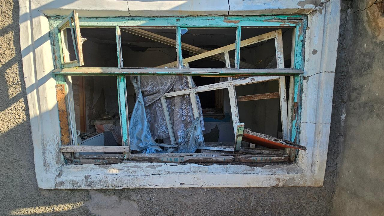 Понівечене вибуховою хвилею вікно приватного будинку в Херсоні з повністю вибитим склом, зламаною дерев'яною рамою та розірваною шторою, що висить усередині кімнати.