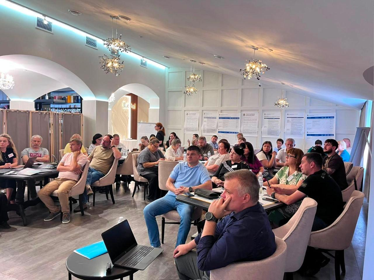 На передньому плані — директор Департаменту розвитку економіки ХОДА Артем Мішин під час презентації проєкту оновленої Стратегії розвитку Херсонської області до 2027 року, де представники громад Бериславського району та «Програми Polaris» обговорюють план відновлення в конференц-залі.