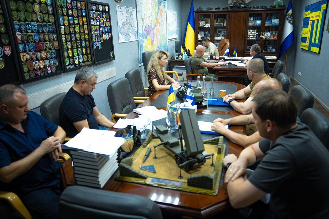 Загальний план засідання Координаційного штабу: на передньому плані праворуч сидить Євген Пісоцький у сірій футболці. У глибині за столом — начальник Херсонської ОВА Олександр Прокудін, праворуч від нього Дмитро Бутрій, ліворуч — Євгеній Ігнатенко. Посередині столу зліва сидить заступниця Ольга Малярчук у сукні в горошок, а майже праворуч навпроти неї — начальник Херсонської МВА Ярослав Шанько в гірчичному поло. На столі розміщено великі макети військової техніки та оборонних споруд.
