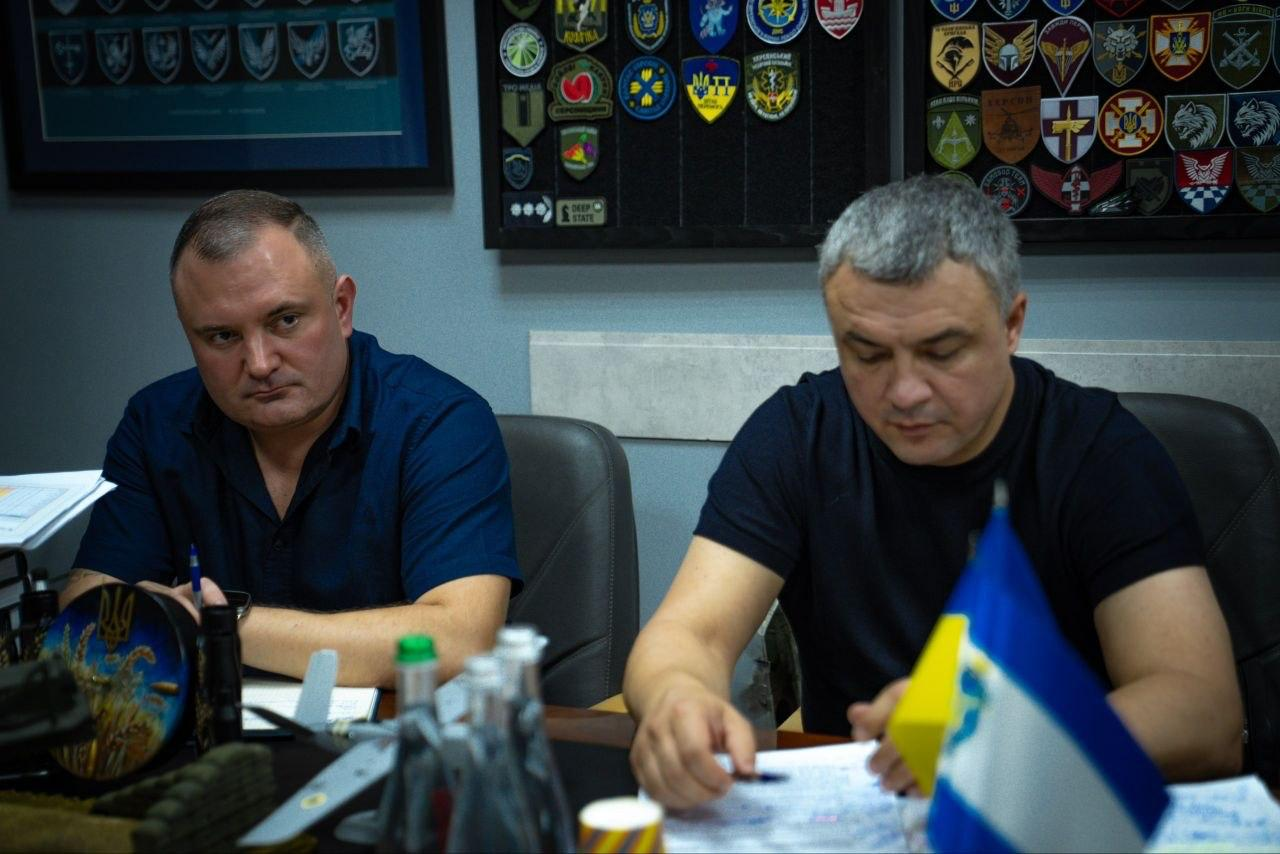 Двоє учасників засідання Координаційного штабу з питань стійкості регіонів та захисту інфраструктури сидять за робочим столом, вивчаючи документи: чоловік ліворуч одягнений у темно-синю сорочку, а чоловік праворуч — у чорну футболку.
