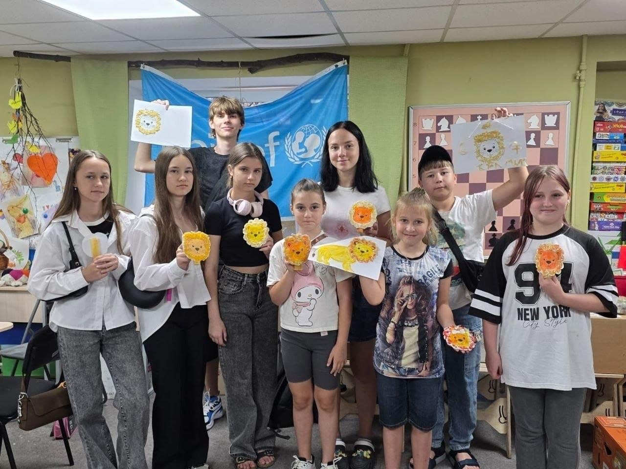Група дітей та підлітків із Херсонщини радісно позує разом у координаційному центрі «Вільні разом» у Дніпрі, тримаючи в руках власноруч виготовлені малюнки та аплікації левів на тлі блакитного прапора ЮНІСЕФ (UNICEF).