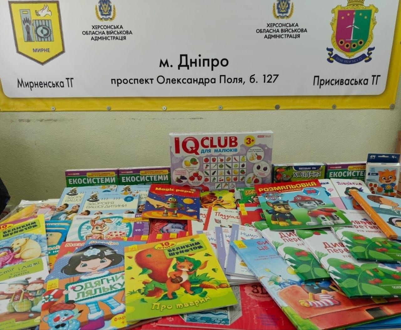 Великий набір дитячих книжок, розмальовок та розвивальних ігор «IQ CLUB», розкладених на столі у гуманітарному хабі в Дніпрі на тлі синьо-жовтого інформаційного стенда Мирненської та Присиваської ТГ.