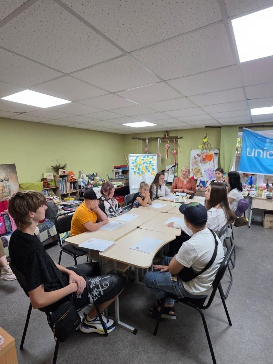 Група дітей та підлітків із Херсонщини сидить за великим столом під час групового заняття в координаційному центрі «Вільні разом» у Дніпрі, а на задньому плані видно блакитний прапор ЮНІСЕФ (UNICEF).