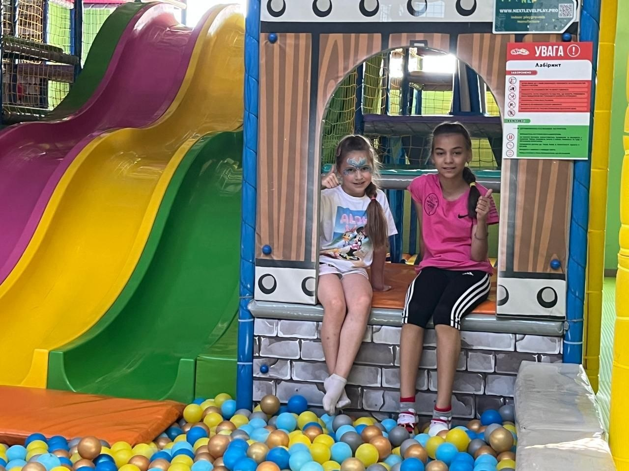 Дві юні переселенки з Херсонщини відпочивають в ігровому лабіринті дитячого центру «Країна мрій» у Хмельницькому. Дівчинка ліворуч — у білій футболці та з аквагримом на обличчі, а дівчинка праворуч — у рожевій футболці, вона радісно посміхається і показує великий палець вгору. Перед ними розташований басейн із синіми, блакитними та жовтими кульками, а поруч — різнокольорові гірки.