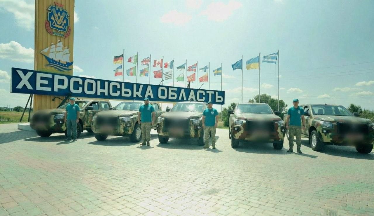 Четверо військових у камуфляжних штанах та бірюзових футболках стоять біля п'яти камуфляжних позашляховиків на в'їзді до Херсонської області. На задньому плані височіє жовта стела з гербом області, кораблем та великим написом «ХЕРСОНСЬКА ОБЛАСТЬ», поруч із якою на флагштоках майорять прапори України та країн-партнерів.