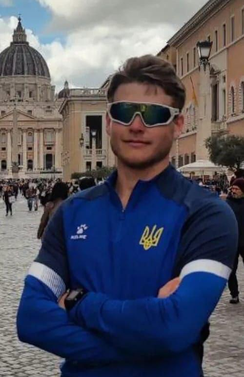 Український веслувальник Олександр Іванов у синій спортивній формі національної збірної України з гербом на грудях. Спортсмен стоїть у сонцезахисних окулярах із дзеркальними лінзами, упевнено схрестивши руки, на тлі міської європейської архітектури.