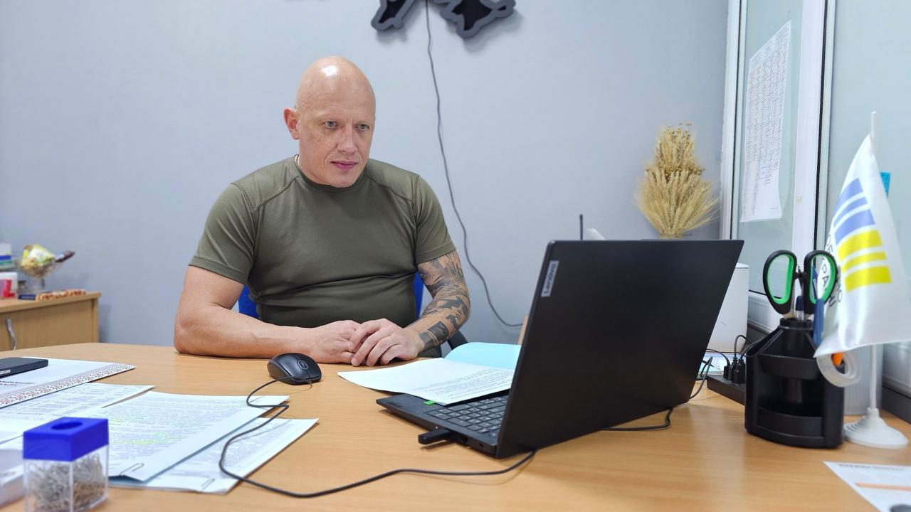 Заступник начальника Херсонської ОВА Євгеній Ігнатенко бере участь у розширеному засіданні колегії Головного управління Держпродспоживслужби в Херсонській області через відеозв'язок. Чоловік у зеленій військовій футболці та з татуюваннями на руці сидить за робочим столом у кабінеті, дивиться в екран чорного ноутбука Lenovo та тримає руки на столі поруч із комп'ютерною мишкою, документами та маленьким прапорцем України.