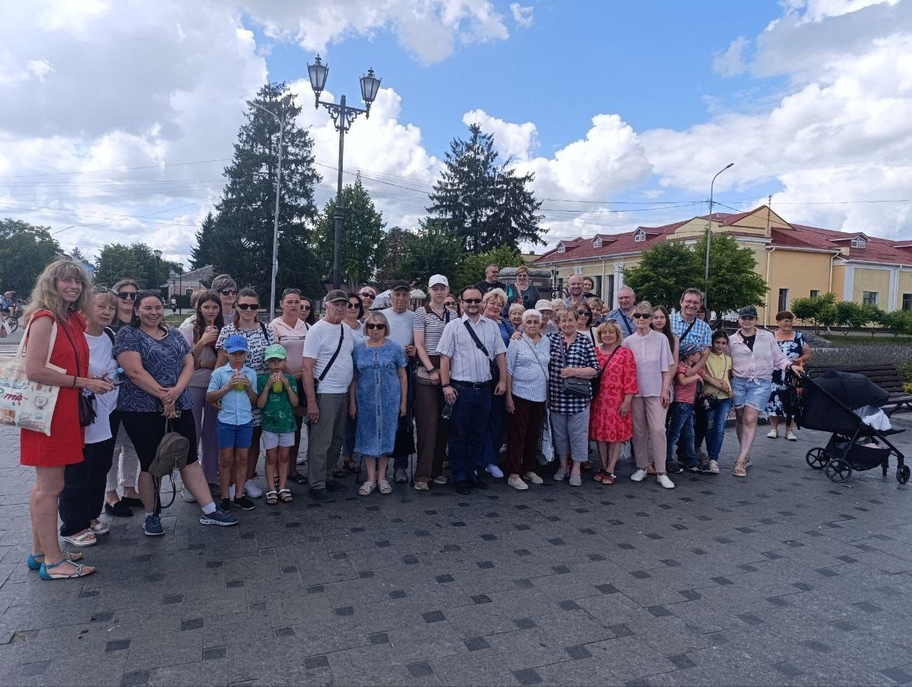 Велика група переселенців із Херсонщини та фахівців координаційного центру «Вільні разом» позує для спільного фото на міській площі Бердичева. У центрі групи стоїть екскурсовод, краєзнавець Олексій Побережний в окулярах та білій сорочці з коротким рукавом. Люди різного віку стоять на брукованій площі на тлі історичних будівель, дерев та літнього неба.