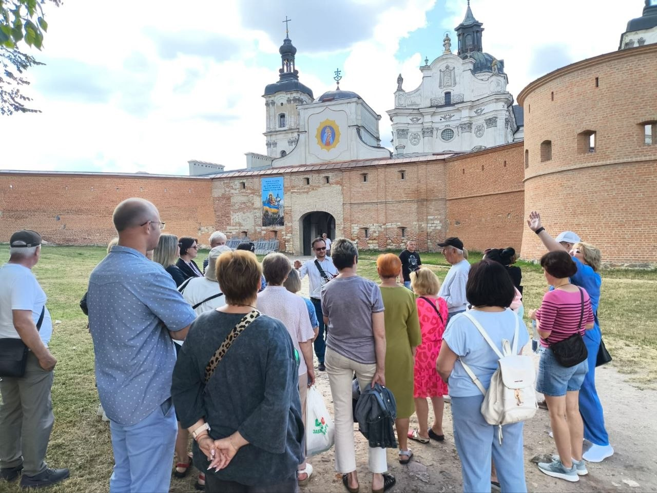 Група переселенців із Херсонщини слухає краєзнавця Олексія Побережного (в окулярах та білій сорочці в центрі) під час екскурсії біля Санктуарію Бердичівської Богородиці (Монастиря Босих Кармелітів). Люди стоять спиною до камери на зеленій галявині перед високим цегляним муром фортеці, за яким височіють вежі та бані старовинного католицького храму під хмарним небом.