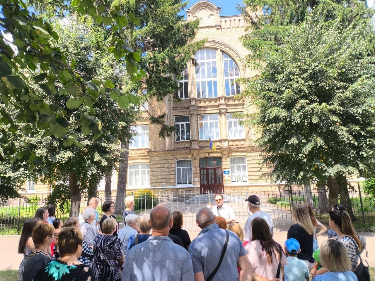 Група переселенців із Херсонщини слухає краєзнавця Олексія Побережного (у білій сорочці та окулярах) під час екскурсії біля історичної будівлі Комерційного училища в Бердичеві. Люди стоять перед металевим парканом навчального закладу, оточеного високими зеленими деревами та ялинами. Сама триповерхова споруда зведена з жовтої цегли, має великі арочні вікна на верхньому поверсі, центральний вхід із дерев'яними дверима та державний прапор України над ними.
