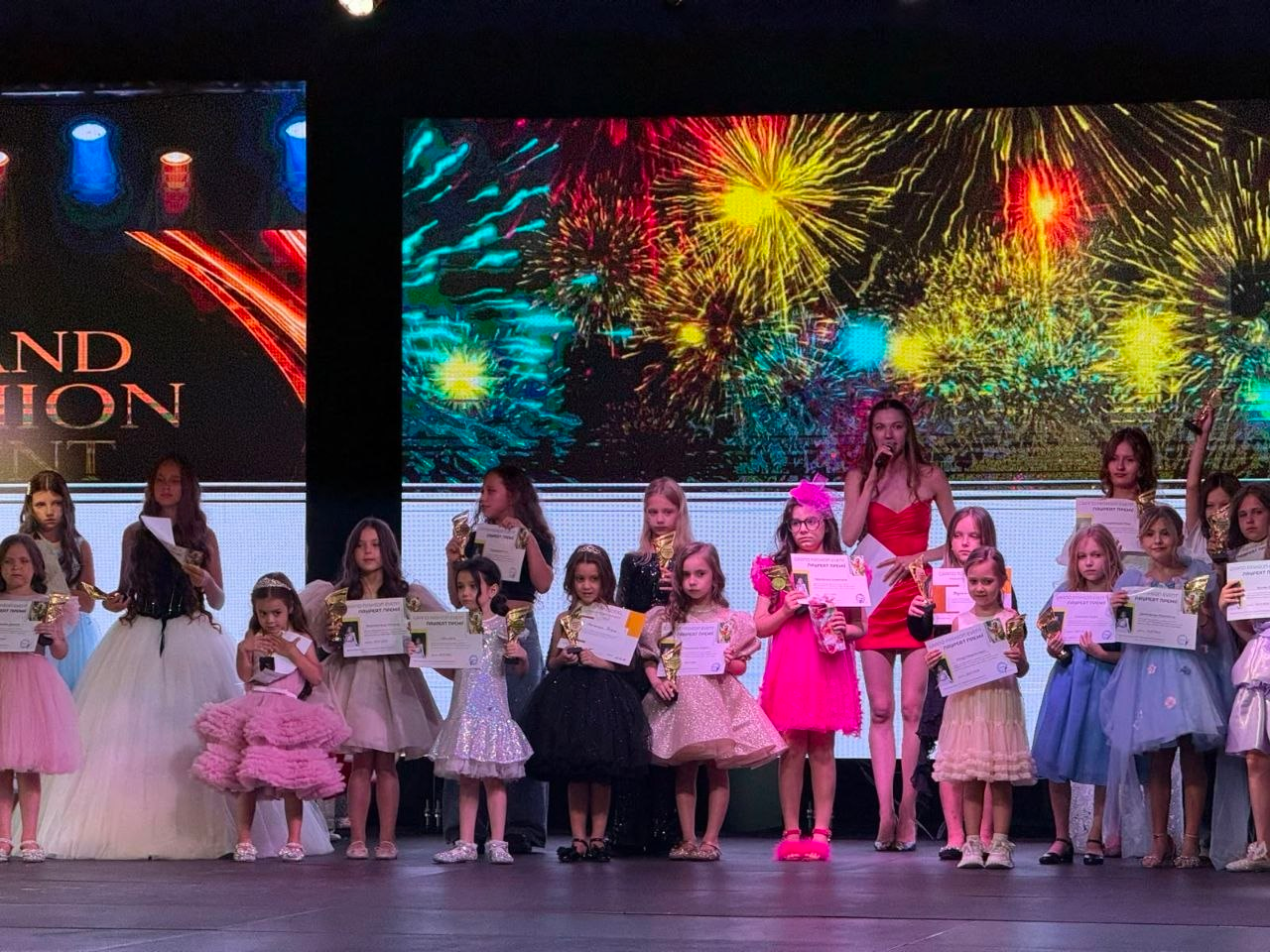 Група юних учасниць модельного показу «Grand Fashion Event» стоїть на сцені під час церемонії нагородження. Дівчатка різного віку в ошатних пишних сукнях тримають у руках дипломи лауреатів та золоті кубки. Позаду них стоїть ведуча в червоній сукні з мікрофоном, а на великому світлодіодному екрані на задньому плані транслюється яскраве зображення святкового салюту.