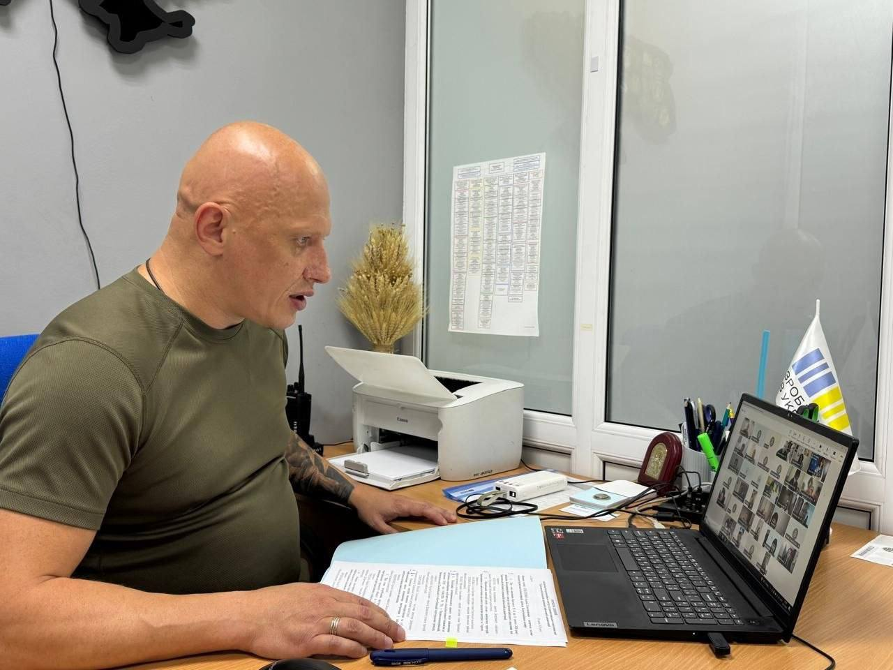 Заступник начальника Херсонської ОВА Євгеній Ігнатенко проводить онлайн-засідання Координаційного центру підтримки цивільного населення. Посадовець у зеленій футболці сидить за робочим столом перед відкритими документами та ноутбуком Lenovo, на екрані якого відображається відеотрансляція робочої зустрічі з багатьма учасниками з громад Херсонщини.