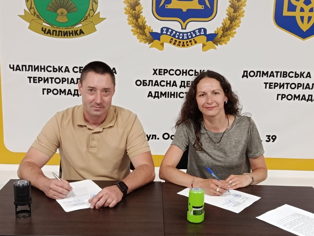 Начальник Чаплинської СВА Генічеського району В'ячеслав Мирошниченко та представниця ГО «ВАЛЕ.О» сидять за столом і підписують меморандум про співпрацю. Вони зафіксовані в момент підписання документів на тлі брендованого банера з гербами та написами Чаплинської громади, Херсонської ОДА та Долматівської громади.