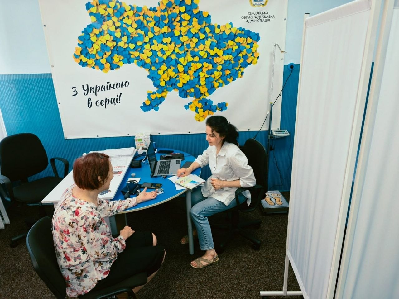Лікарка-терапевтка у білому халаті консультує жінку-переселенку за синім робочим столом у центрі «Вільні разом». На столі стоїть відкритий ноутбук, лежать медичні картки та тонометр, а на задньому плані висить великий плакат Херсонської ОДА у формі карти України з жовто-блакитних сердець та написом «З Україною в серці!».