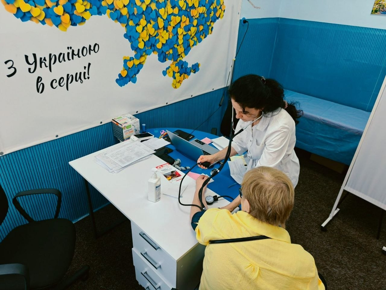 Лікарка-терапевтка у білому медичному халаті вимірює артеріальний тиск жінці в жовтій сорочці за робочим столом у центрі «Вільні разом». Лікарка слухає пульс за допомогою стетоскопа, а над ними на стіні висить плакат із частиною карти України з жовто-блакитних сердець та написом «З Україною в серці!».