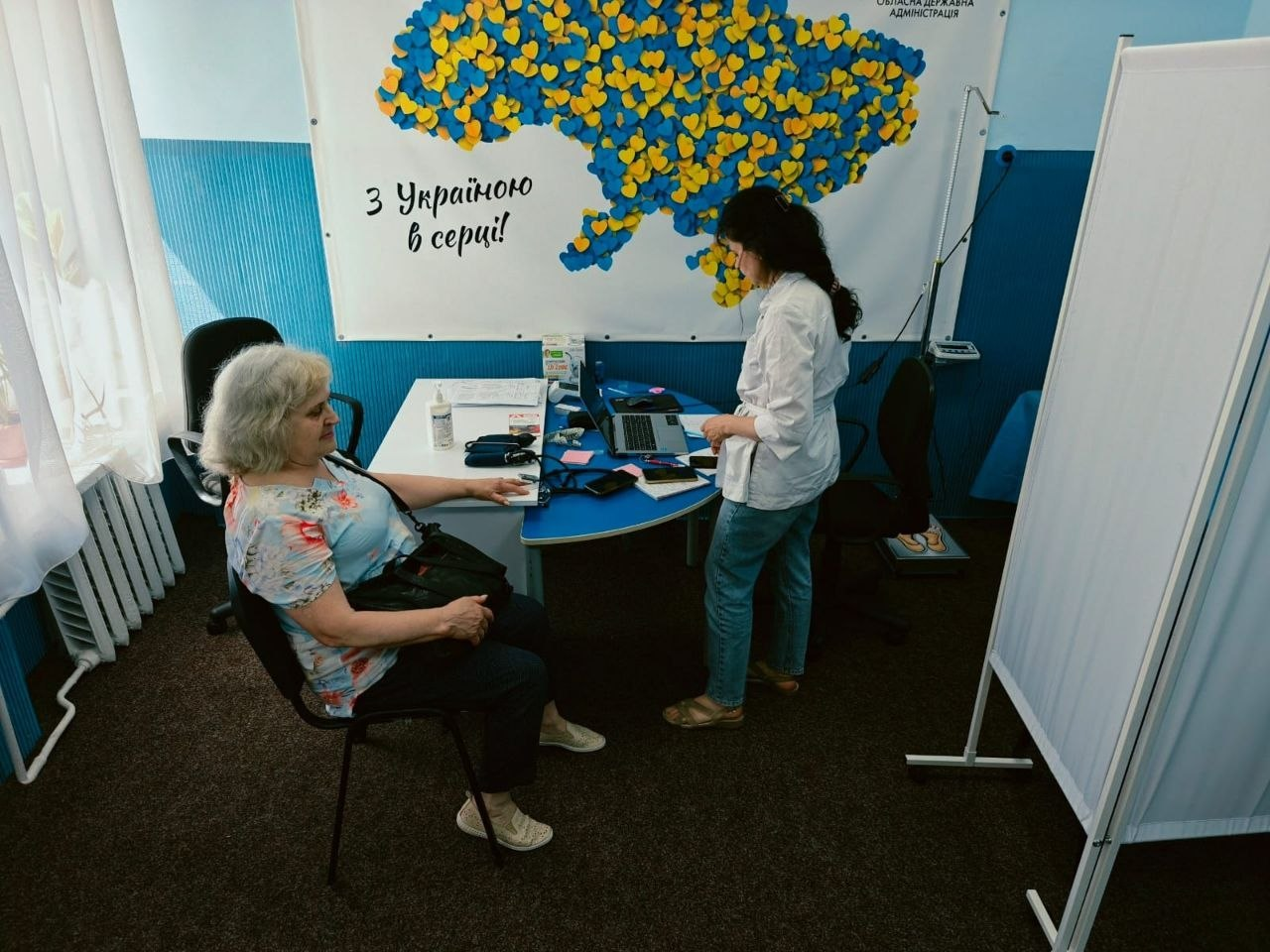 Лікарка-терапевтка у білому медичному халаті стоїть біля робочого столу та консультує сивочолу жінку-переселенку в блакитній футболці, яка сидить на стільці поруч. Прийом проходить у медичному кабінеті центру «Вільні разом» на тлі великого плаката з картою України та написом «З Україною в серці!».
