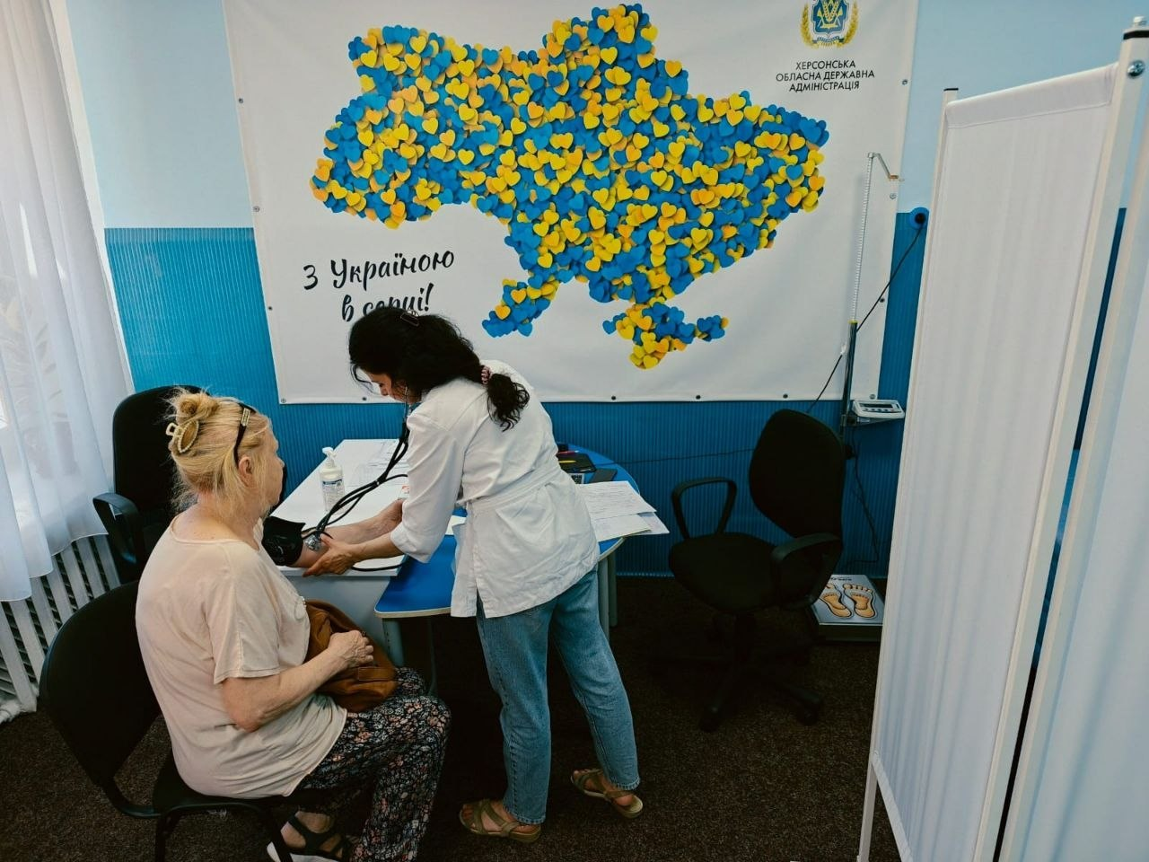 Лікарка-терапевтка у білому медичному халаті схилилася над літньою пацієнткою, щоб виміряти їй артеріальний тиск за допомогою тонометра. Прийом проходить у кабінеті центру «Вільні разом» на тлі великого банера з гербом Херсонської ОДА, картою України з жовто-блакитних сердець та написом «З Україною в серці!».