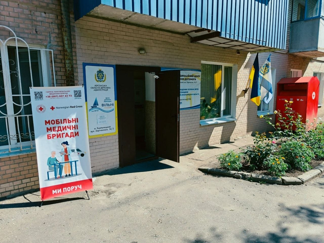 Вхід до Координаційного центру допомоги переселенцям з Херсонщини у Хмельницькому. Біля відчинених дверей цегляної будівлі встановлено банер «Мобільні медичні бригади» від Червоного Хреста та таблички з гербом Херсонської ОДА й логотипом центру «Вільні разом», а праворуч на стіні майорять прапори України та Херсонської області.