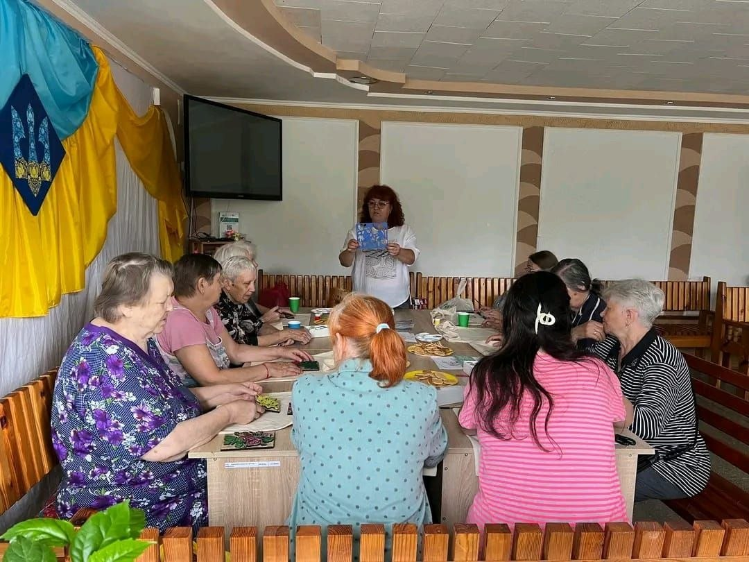 Учасники творчого заняття сидять за великим столом у Центрі життєстійкості, а ведуча стоїть на чолі столу та демонструє зразок виробу. На столі розкладені екосумки-шопери для розпису та матеріали для творчості, а ліворуч на стіні висить великий прапор України з гербом.