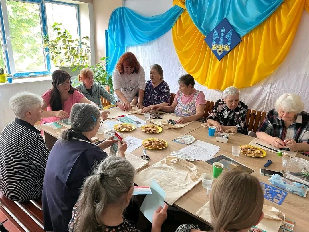 Учасниці творчого майстер-класу в Центрі життєстійкості розписують екосумки-шопери за великим столом. Ведуча схилилася над однією з учасниць, допомагаючи їй із роботою. На столі розкладені тканинні сумки, палітри з фарбами, трафарети та тарілки з печивом, а на стіні позаду висить великий синьо-жовтий прапор із гербом України.