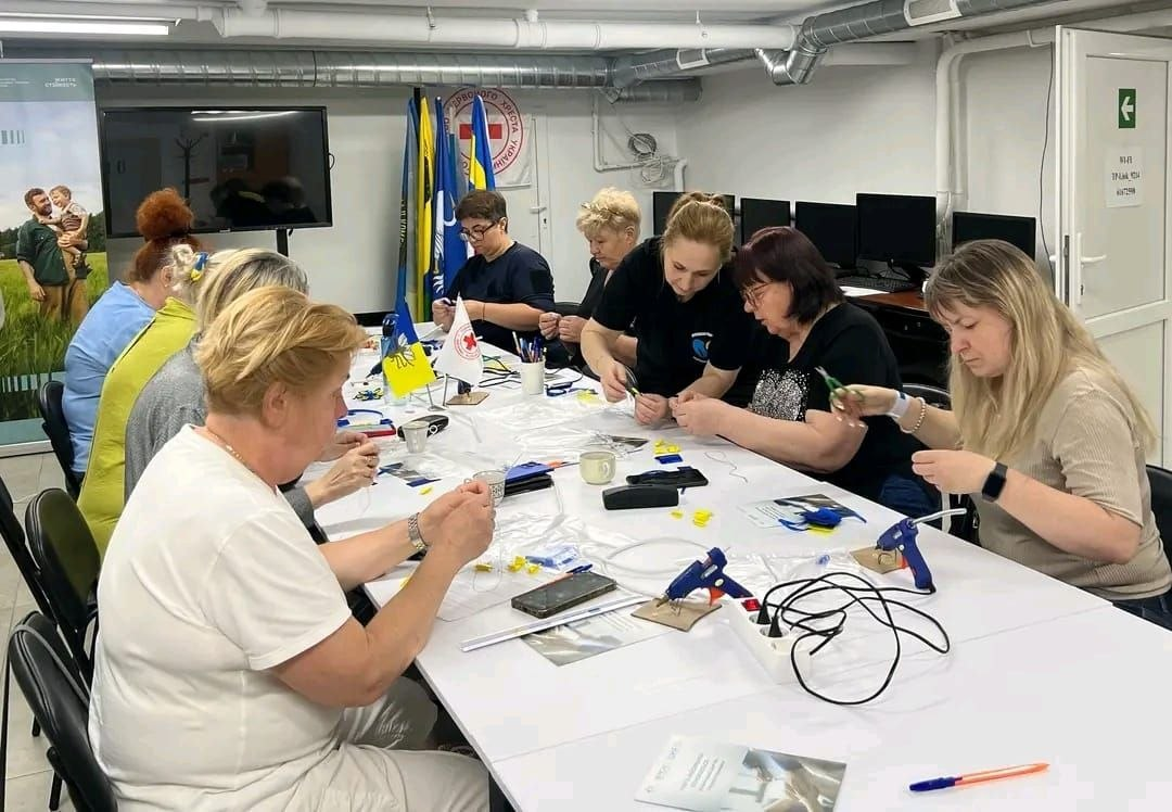 Учасниці майстер-класу в Центрі життєстійкості створюють патріотичні брошки за великим білим столом. На столі розкладені матеріали для творчості, клейові пістолети, а також стоять маленькі прапорці України та Червоного Хреста. На задньому плані видно символіку Товариства Червоного Хреста України та комп'ютерну техніку.