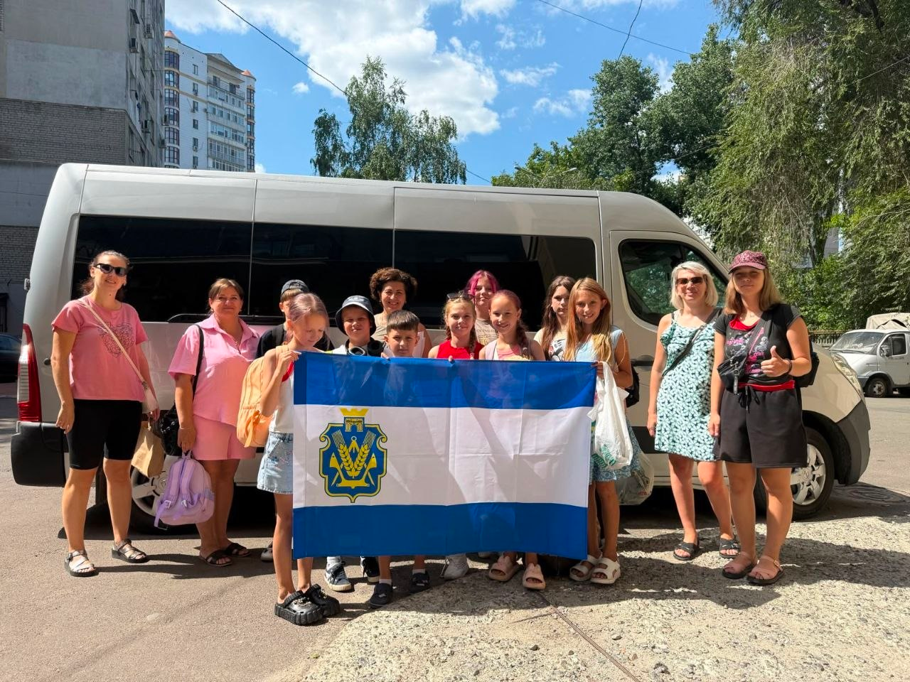 Сьома група дітей-переселенців із Херсонщини разом із дорослими стоїть перед білим мікроавтобусом і тримає прапор Херсонської області перед виїздом на триденний відпочинок біля Хаджибейського лиману на Одещині. Поїздка організована завдяки співпраці координаційного центру «Вільні разом» в Одесі та БФ «Шлях до Дому».