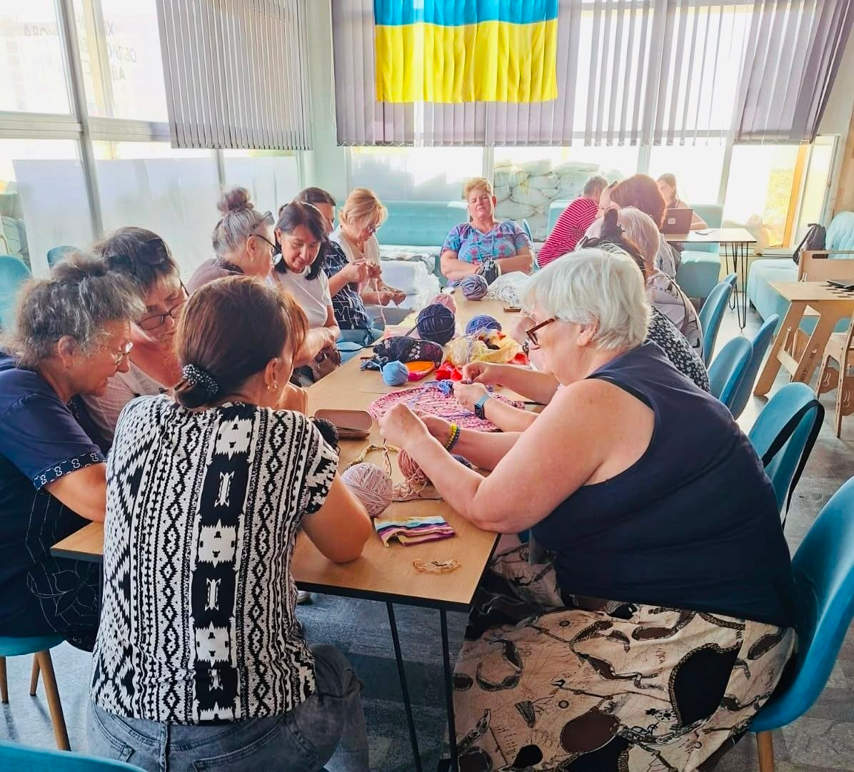 Велика група жінок-переселенок із Херсонщини сидить за довгим столом у світлій кімнаті з українським прапором на вікні. Вони зосереджено обговорюють процес рукоділля та разом плетуть круглі килимки гачками з кольорової пряжі під час заняття гуртка «Коло тепла» в координаційному центрі в Миколаєві.