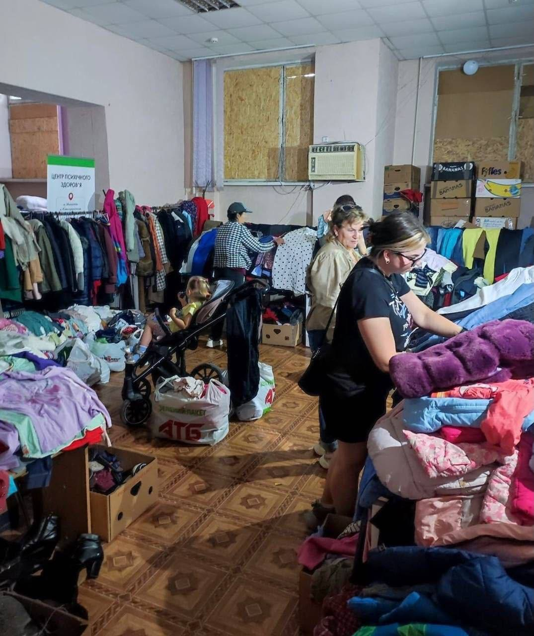 Люди обирають речі у куточку безоплатного одягу від координаційного центру «Вільні разом» у Миколаєві. На тлі розвішаних та складених речей видно дитячий візок, коробки та табличку «Центр психічного здоров'я».