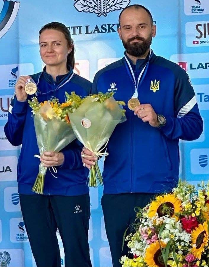 Заслужена майстриня спорту України Галина Авраменко та її колега по збірній у синій формі тримають завойовані медалі й букети квітів під час нагородження на Чемпіонаті світу з кульової стрільби.