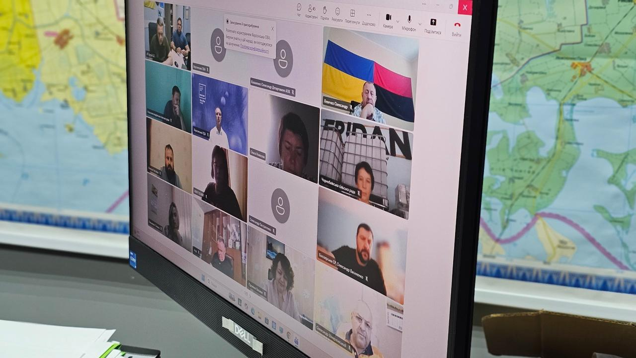 На екрані комп'ютера відображено вікна програми для відеоконференцій Microsoft Teams, де на зв'язку перебувають керівники місцевих адміністрацій, представники Головного управління Державної податкової служби та Головного управління Держгеокадастру у Херсонській області, які обговорюють проблемні питання аграрного бізнесу регіону. На тлі за монітором видно настінну мапу.