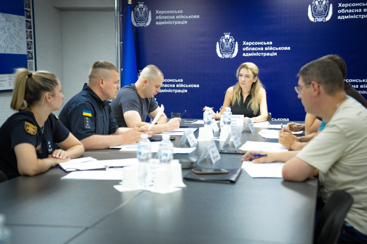 Заступник начальника Херсонської ОВА Ольга Малярчук проводить нараду щодо готовності укриттів. Ліворуч за столом сидить начальник Херсонської МВА Ярослав Шанько, а праворуч — директор Департаменту з питань цивільного захисту та оборонної роботи ХОДА Євген Пісоцький, частково закритий іншим учасником наради. Також присутні представники ДСНС та Нацполіції на тлі синього банера з логотипом ХОВА.