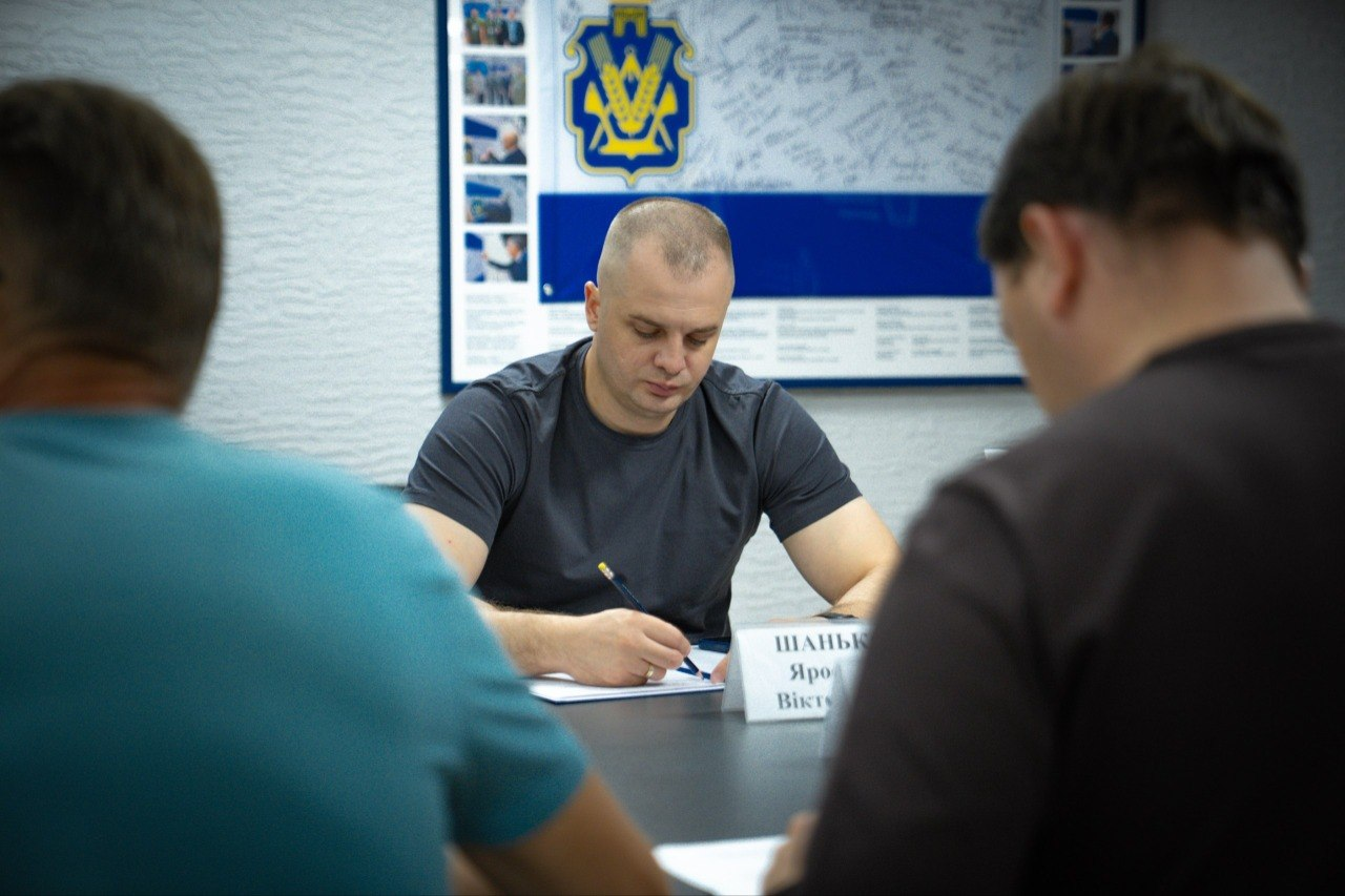 Начальник Херсонської МВА Ярослав Шанько сидить за столом під час наради щодо готовності укриттів та робить записи в документах. На передньому плані спиною до камери сидять учасники зустрічі: праворуч — директор Департаменту з питань цивільного захисту та оборонної роботи ХОДА Євген Пісоцький, ліворуч — заступник директора Департаменту – начальник управління Володимир Бєляєв. На задньому плані — інформаційний стенд із символікою.
