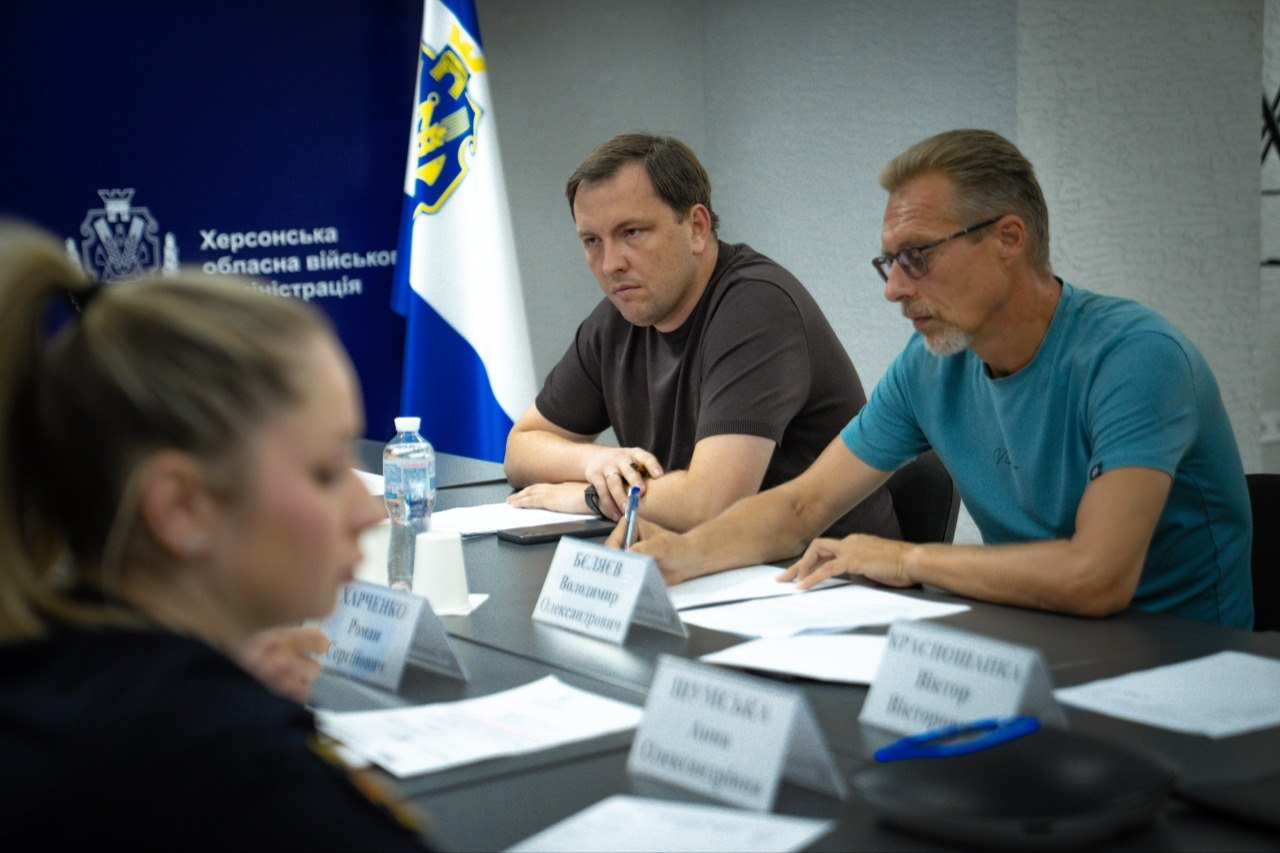 Учасники наради щодо готовності укриттів сидять за робочим столом: ліворуч — директор Департаменту з питань цивільного захисту та оборонної роботи ХОДА Євген Пісоцький, праворуч — заступник директора Департаменту – начальник управління Володимир Бєляєв. На передньому плані спиною до камери сидить представниця Нацполіції, а на задньому плані видно Державний Прапор України та синій банер із логотипом Херсонської ОВА.